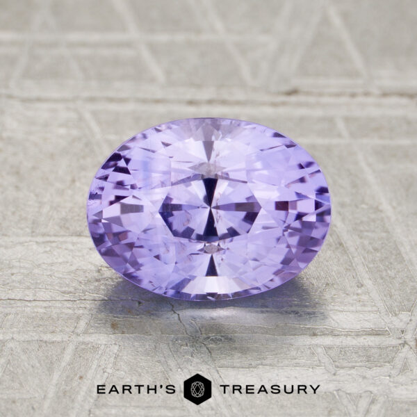1.81-Carat Ceylon Sapphire A 1.81-carat faceted oval Ceylon sapphire with a lavender hue rests on a textured light gray surface, with the "Earth’s Treasury" logo displayed at the bottom.