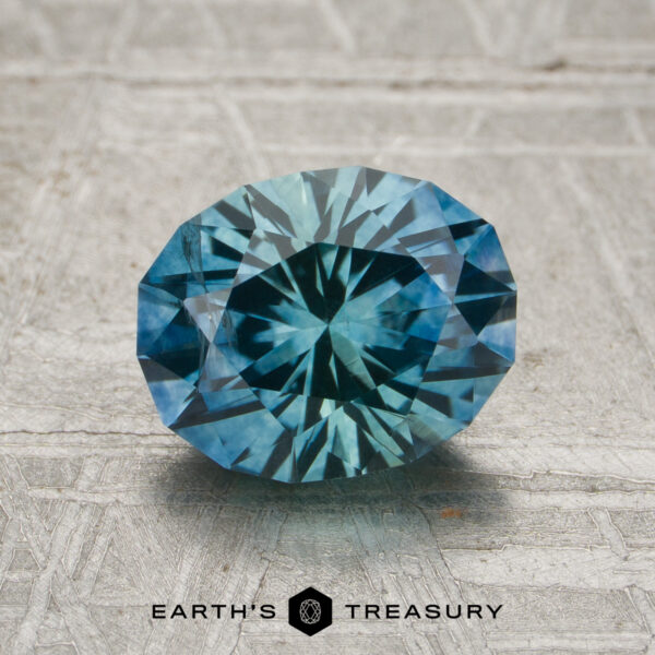 1.95-Carat Aqua-Blue Particolored Montana Sapphire (Heated) A 1.95-carat aqua-blue particolored Montana sapphire (heated) rests on a textured gray surface, with the "EARTH'S TREASURY" text and logo displayed beneath it.
