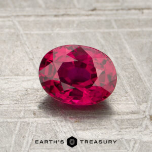 A 1.17-carat heated Mozambique ruby with an oval cut is displayed on a textured metallic surface, accompanied by the "EARTH'S TREASURY" text and a hexagonal logo below.