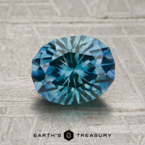 A 1.94-carat aqua-blue particolored Montana sapphire (heated) with intricate faceting is displayed on a textured gray surface, with the Earth’s Treasury logo below.