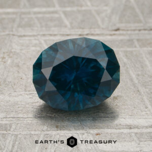 A 1.93-carat Peacock Blue Montana Sapphire (heated) is displayed on a textured gray surface, with the "EARTH'S TREASURY" text and logo below it.