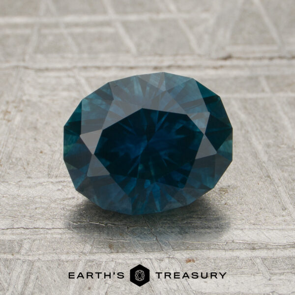 1.93-Carat Peacock Blue Montana Sapphire (Heated) A 1.93-carat Peacock Blue Montana Sapphire (heated) is displayed on a textured gray surface, with the "EARTH'S TREASURY" text and logo below it.