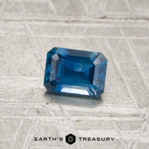 A rectangular, deep blue, faceted 0.69-carat Montana sapphire (heated) rests on a textured silver surface. The text "EARTH'S TREASURY" appears at the bottom of the image.