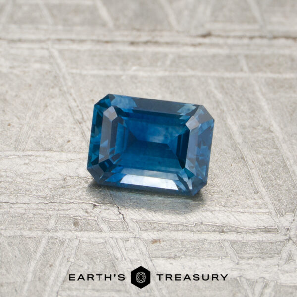 0.69-Carat Deep Blue Montana Sapphire (Heated) A rectangular, deep blue, faceted 0.69-carat Montana sapphire (heated) rests on a textured silver surface. The text "EARTH'S TREASURY" appears at the bottom of the image.