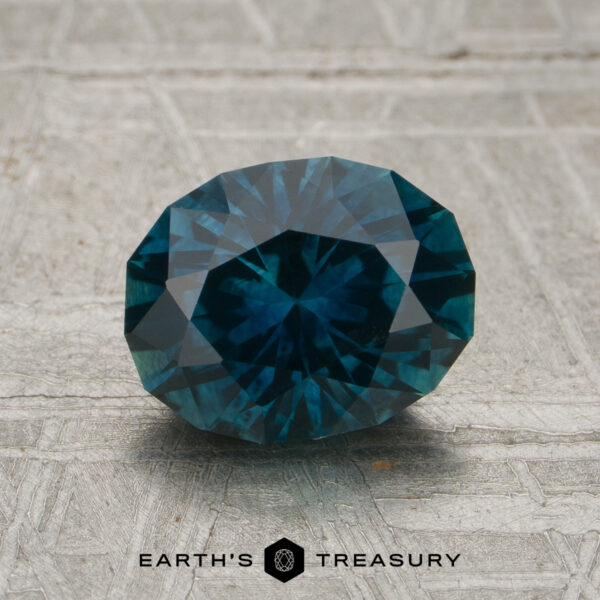 1.93-Carat Peacock Montana Sapphire (Heated) A 1.93-carat Peacock Montana Sapphire (heated), cut in a faceted oval shape with vibrant blue-green hues, rests on a textured gray surface featuring the "EARTH'S TREASURY" text and a hexagonal logo below the gemstone.
