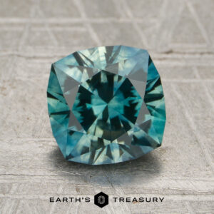 A 2.17-carat aqua green-teal particolored Montana sapphire (heated) with a cushion cut is displayed on a gray textured surface. "Earth's Treasury" is printed below the gem.