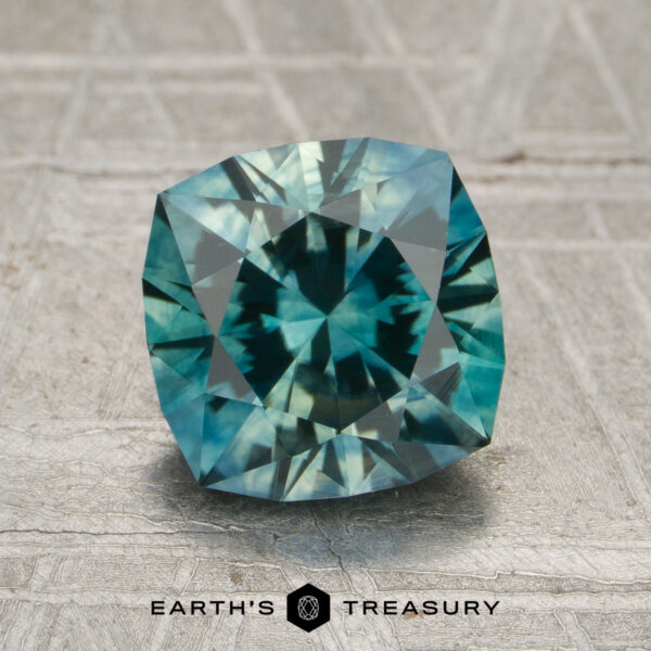 2.17-Carat Teal-Green Particolored Montana Sapphire (Heated) A 2.17-carat aqua green-teal particolored Montana sapphire (heated) with a cushion cut is displayed on a gray textured surface. "Earth's Treasury" is printed below the gem.