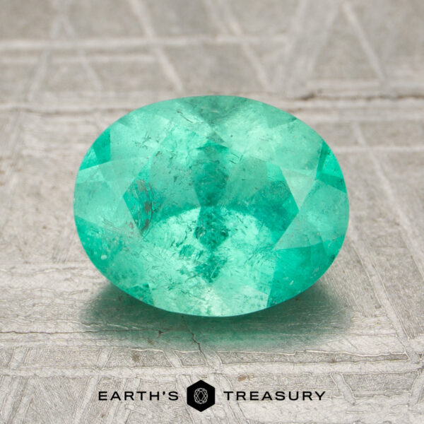 1.73-Carat Bright Green Colombian Emerald A 1.73-carat bright green Colombian emerald, oval-cut, is displayed on a textured metallic surface with the Earth’s Treasury logo at the bottom.
