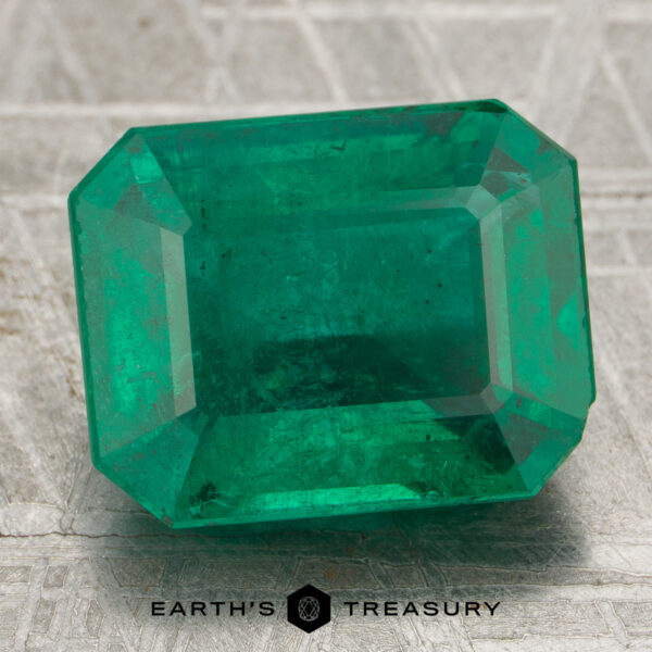 The 6.52-carat Rich Green Zambian Emerald, cut in a rectangular shape, is displayed on a textured gray surface with the Earth's Treasury logo at the bottom.