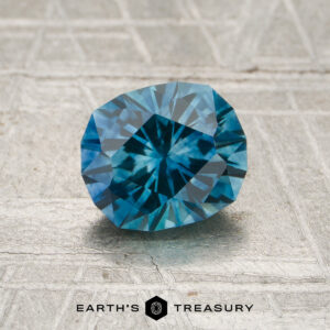 A 1.43-carat teal blue Montana sapphire (heated) is displayed on a textured gray surface with the text "EARTH'S TREASURY" at the bottom.