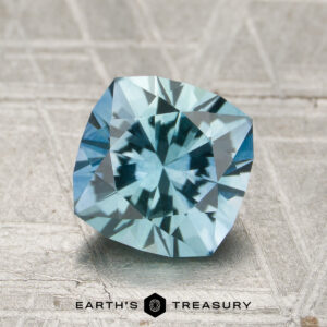 A 1.91-carat, cushion-cut aqua blue Montana sapphire (heated) rests on a textured surface with the logo and text "EARTH'S TREASURY" visible at the bottom of the image.