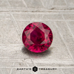 A 0.66-carat round-cut red Mozambique ruby (heated) is displayed on a textured gray surface with the Earth's Treasury logo at the bottom.