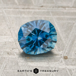 A 1.38-carat teal blue Montana sapphire (heated) is showcased on a textured surface, with the "EARTH'S TREASURY" logo and text visible at the bottom.