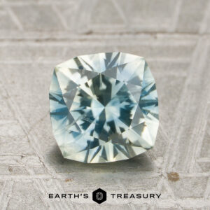 A faceted, cushion-cut 1.85-carat pale blue-yellow particolored Montana sapphire (heated) rests on a textured surface. The "Earth's Treasury" logo and text are displayed at the bottom of the image.