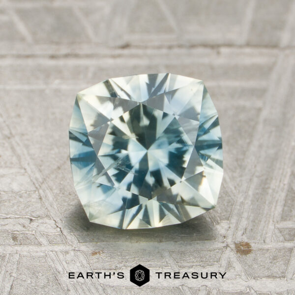 1.85-Carat Pale Blue-Yellow Particolored Montana Sapphire (Heated) A faceted, cushion-cut 1.85-carat pale blue-yellow particolored Montana sapphire (heated) rests on a textured surface. The "Earth's Treasury" logo and text are displayed at the bottom of the image.