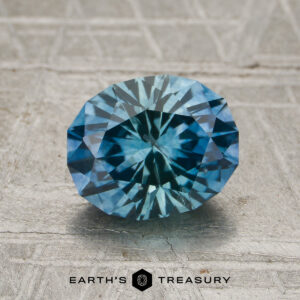 A 1.90-carat teal-blue particolored Montana sapphire (heated) rests on a textured gray surface, with the words "Earth's Treasury" and a hexagon logo displayed below.