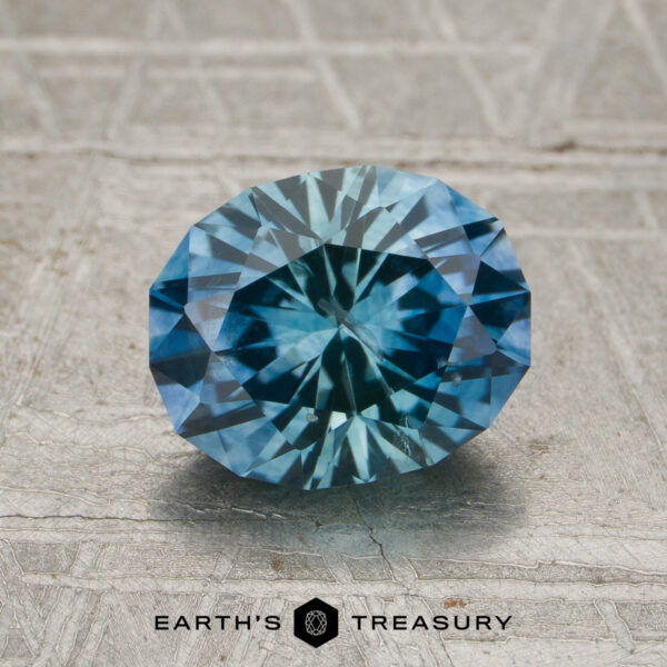 1.90-Carat Teal-Blue Particolored Montana Sapphire (Heated) A 1.90-carat teal-blue particolored Montana sapphire (heated) rests on a textured gray surface, with the words "Earth's Treasury" and a hexagon logo displayed below.
