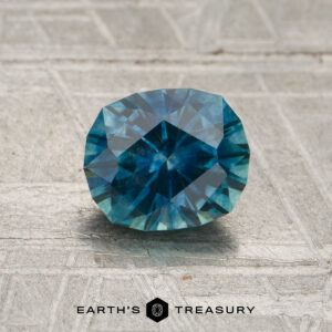A 1.32-carat teal Montana sapphire (heated) rests on a textured gray surface, with the "EARTH'S TREASURY" logo displayed below.