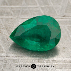 A pear-shaped 2.12-carat rich green Zambian emerald gemstone rests on a textured surface, with the logo and text "Earth's Treasury" visible below.