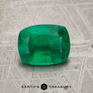 A 2.05-carat rich green Zambian emerald is displayed on a textured, patterned surface with the text "Earth's Treasury" below it.