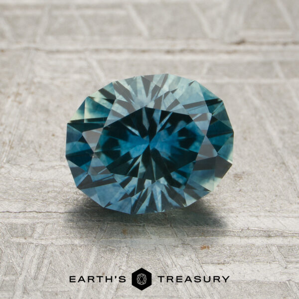 1.86-Carat Teal Montana Sapphire (Heated) A 1.86-carat teal Montana sapphire (heated), expertly oval-cut with intricate facets, is displayed on a textured, light-colored surface. The text "EARTH'S TREASURY" appears at the bottom.