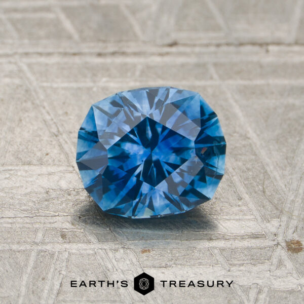 A 1.20-carat rich blue Montana sapphire (heated), featuring a faceted round cut, is displayed on a textured light gray surface with the "Earth's Treasury" text and logo at the bottom.