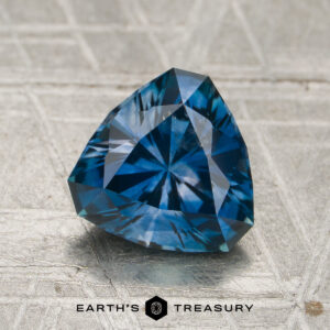 A 1.81-carat rich blue Montana sapphire (heated) with a triangular shape rests on a textured surface, with the "Earth's Treasury" text visible at the bottom of the image.