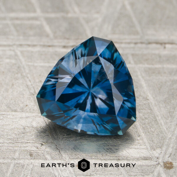 1.81-Carat Deep Rich Blue Montana Sapphire (Heated) A 1.81-carat rich blue Montana sapphire (heated) with a triangular shape rests on a textured surface, with the "Earth's Treasury" text visible at the bottom of the image.