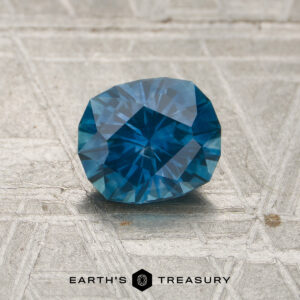 A 1.13-carat Peacock Blue Montana Sapphire (heated) rests on a textured, metallic surface adorned with the "EARTH'S TREASURY" text and a hexagonal logo below it.