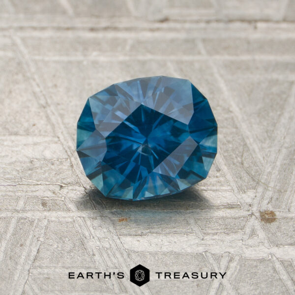 1.13-Carat Peacock Blue Montana Sapphire (Heated) A 1.13-carat Peacock Blue Montana Sapphire (heated) rests on a textured, metallic surface adorned with the "EARTH'S TREASURY" text and a hexagonal logo below it.