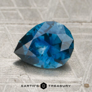 A 1.74-carat rich teal blue Montana sapphire (heated) with a faceted cut rests on a textured, silvery surface featuring the "EARTH'S TREASURY" name and a hexagonal logo below.