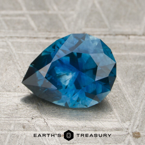 1.74-Carat Rich Teal Blue Montana Sapphire (Heated) A 1.74-carat rich teal blue Montana sapphire (heated) with a faceted cut rests on a textured, silvery surface featuring the "EARTH'S TREASURY" name and a hexagonal logo below.