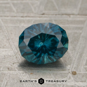 A 1.83-carat Peacock Montana Sapphire (heated), with a faceted teal blue hue, rests on a textured gray surface alongside the "EARTH'S TREASURY" text and hexagon logo at the bottom.