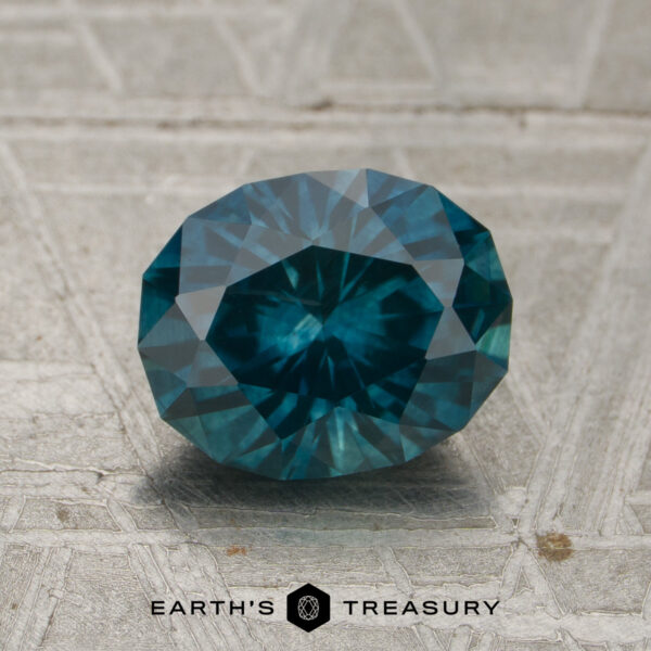 1.83-Carat Peacock Montana Sapphire (Heated) A 1.83-carat Peacock Montana Sapphire (heated), with a faceted teal blue hue, rests on a textured gray surface alongside the "EARTH'S TREASURY" text and hexagon logo at the bottom.