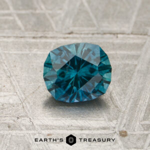 A 1.03-carat rich teal Montana sapphire (heated) rests on a textured surface beside the text "Earth’s Treasury" and a hexagon logo.