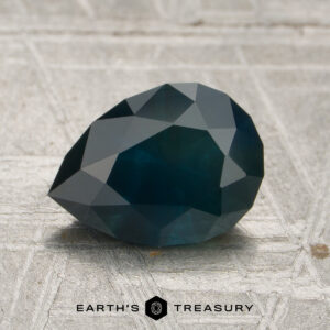 A 1.69-carat Montana Sapphire (heated) with a dark blue, faceted appearance is displayed on a textured surface featuring the Earth's Treasury logo and text at the bottom.