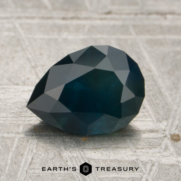 1.69-Carat Near-Black Midnight Blue Montana Sapphire (Heated) A 1.69-carat Montana Sapphire (heated) with a dark blue, faceted appearance is displayed on a textured surface featuring the Earth's Treasury logo and text at the bottom.