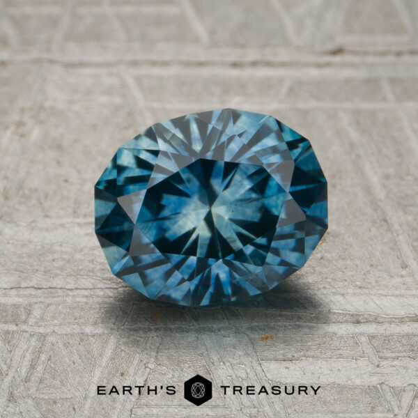 A 1.81-carat medium blue Montana sapphire (heated) is displayed on a textured surface with the words "EARTH'S TREASURY" and a logo beneath it.