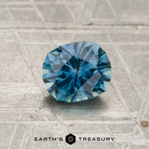 A 1.03-carat teal blue Montana sapphire (heated) rests on a textured gray surface, with the Earth's Treasury logo appearing at the bottom of the image.
