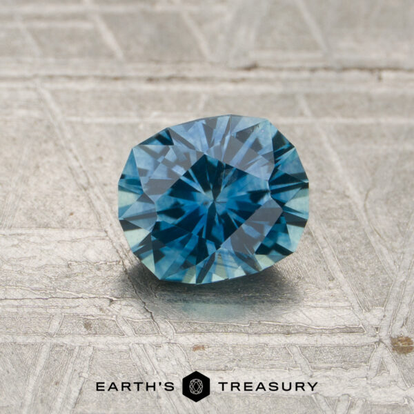 1.03-Carat Teal Blue Montana Sapphire (Heated) A 1.03-carat teal blue Montana sapphire (heated) rests on a textured gray surface, with the Earth's Treasury logo appearing at the bottom of the image.