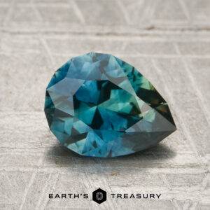 A 1.67-carat blue-green particolored Montana sapphire (heated), faceted in a pear shape, rests on a textured gray surface with the text "EARTH'S TREASURY" visible below the gem.