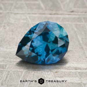 A 1.52-carat teal blue Montana sapphire (heated) rests on a light textured surface, with the text "Earth's Treasury" and a hexagon logo displayed below it.