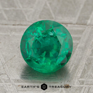 A 1.65-carat rich green Zambian emerald, precision round-cut, rests on a textured gray surface with the text "Earth's Treasury" at the bottom.