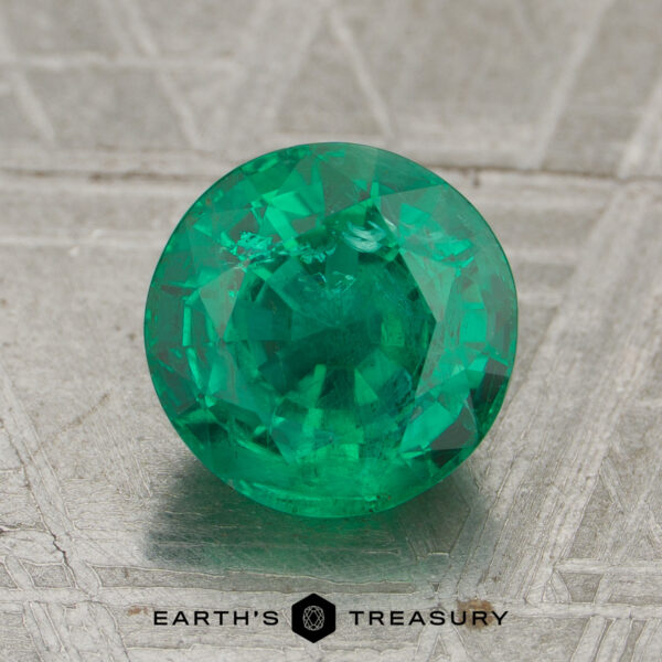 1.65-Carat Rich Green Zambian Emerald A 1.65-carat rich green Zambian emerald, precision round-cut, rests on a textured gray surface with the text "Earth's Treasury" at the bottom.