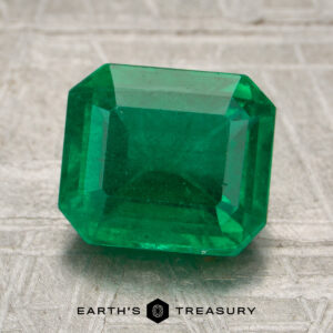 A 2.56-carat rich green Zambian emerald, cut in a rectangular shape, is displayed on a textured surface with the Earth’s Treasury logo beneath it.