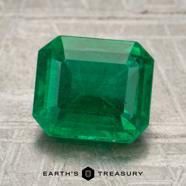 2.56-Carat Rich Green Zambian Emerald A 2.56-carat rich green Zambian emerald, cut in a rectangular shape, is displayed on a textured surface with the Earth’s Treasury logo beneath it.