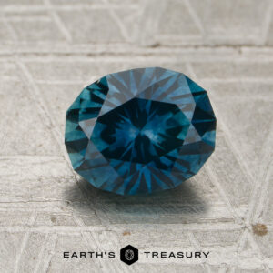 A 1.78-carat Peacock Blue Montana Sapphire (heated) is displayed on a textured gray surface, with "EARTH'S TREASURY" and a small logo featured at the bottom.