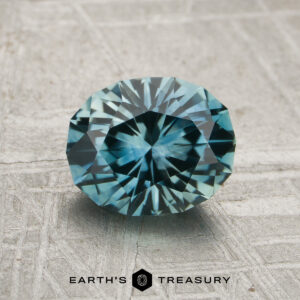 A 1.75-carat aqua blue-green Montana sapphire (heated), cut with round facets, is displayed on a textured gray surface featuring the "Earth's Treasury" logo at the bottom.
