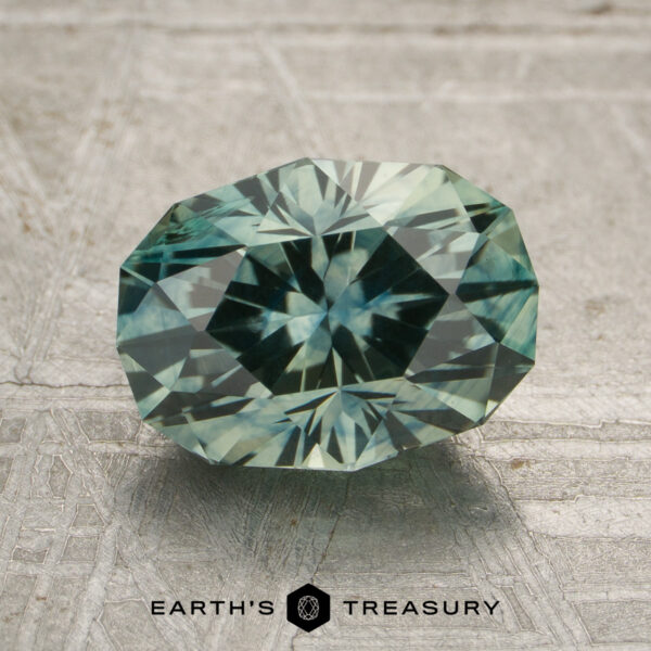 2.30-Carat Montana Sapphire (Heated) A 2.30-carat heated Montana sapphire with green-blue facets is displayed on a textured surface, with the "EARTH'S TREASURY" text and logo beneath it.
