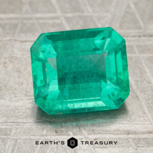A 2.45-carat bright green Brazilian emerald with an emerald cut sits on a textured surface. The words "EARTH'S TREASURY" and a hexagon logo appear at the bottom of the image.